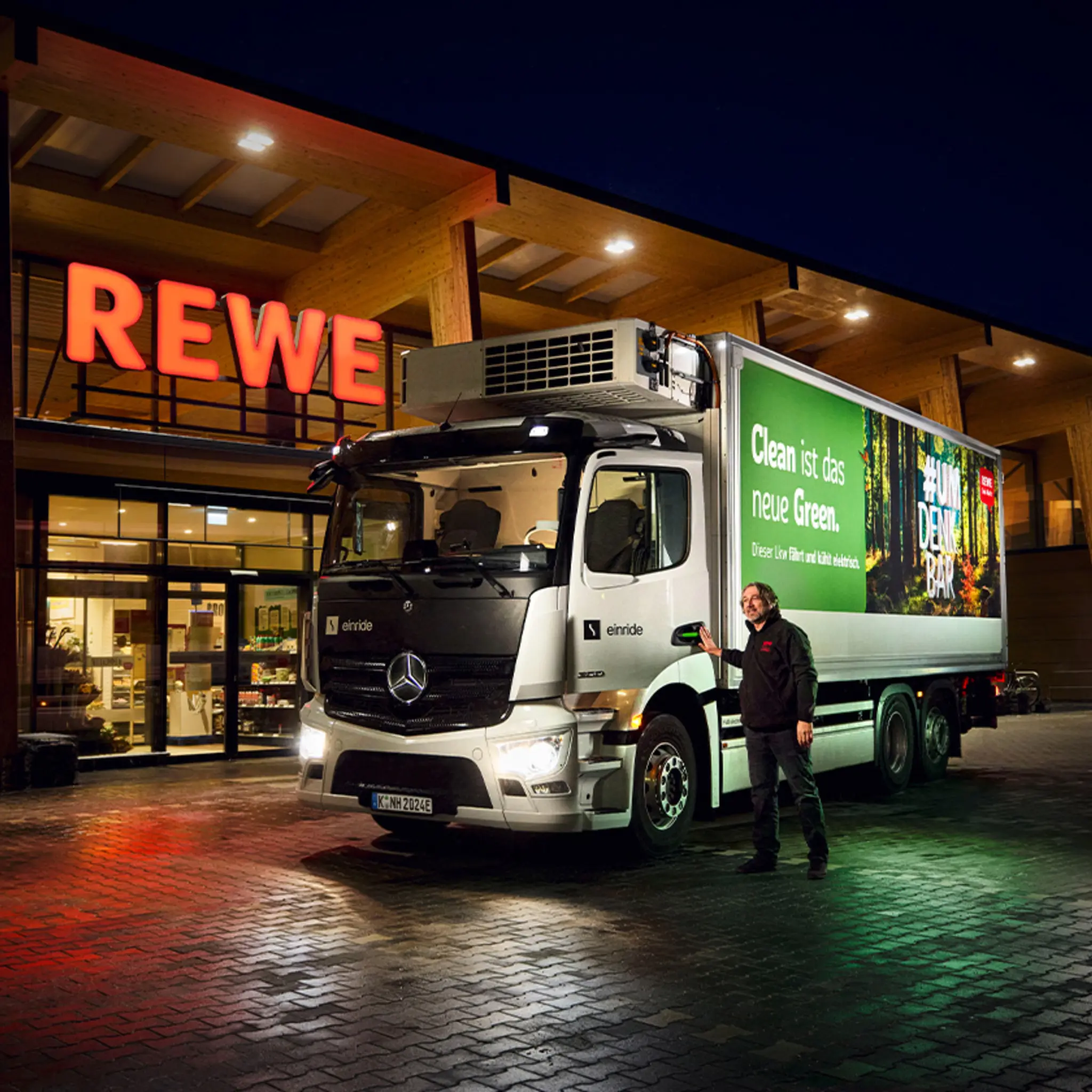Ein mit Grünstrom beleuchteter REWE Markt bei Dunkelheit. Davor hält ein emissionsärmerer REWE E-Lkw.