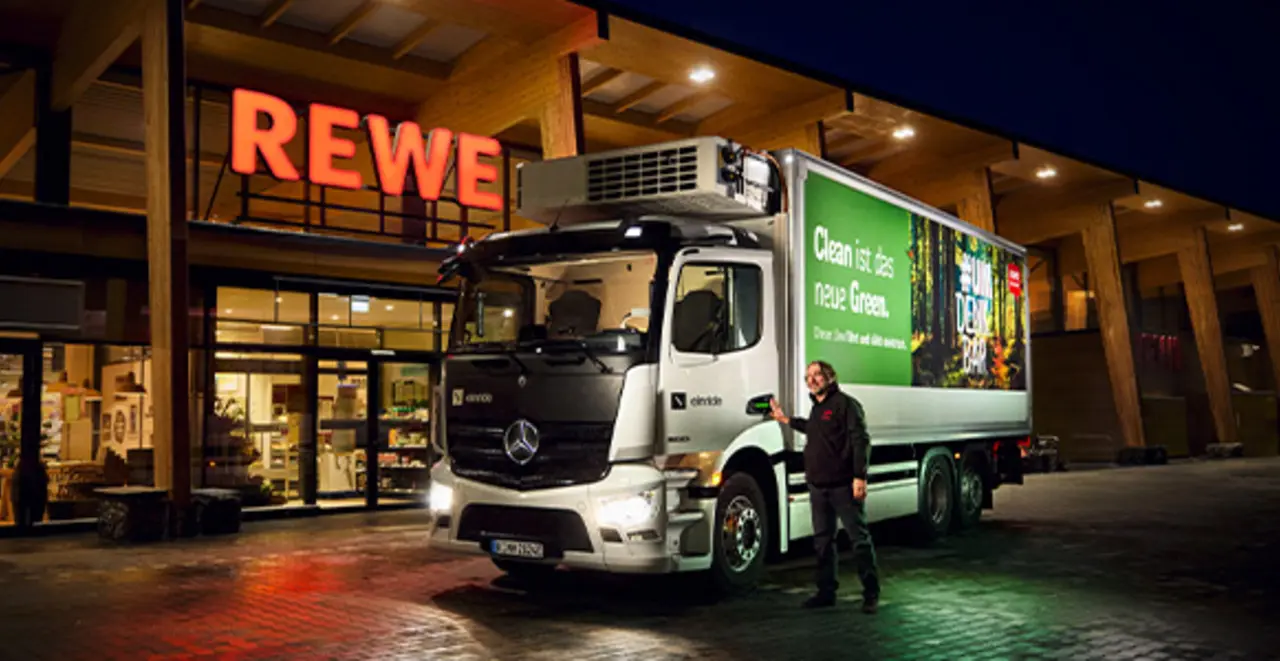 Ein mit Grünstrom beleuchteter REWE Markt bei Dunkelheit. Davor hält ein emissionsärmerer REWE E-Lkw.