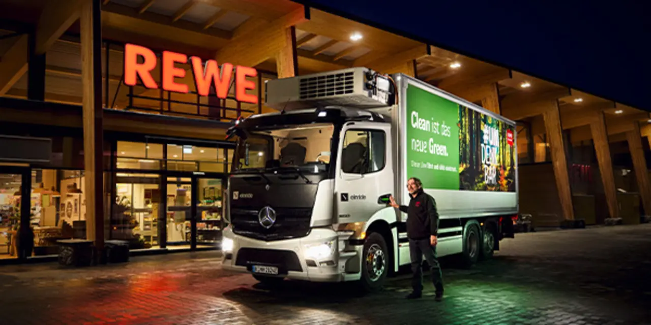 Ein mit Grünstrom beleuchteter REWE Markt bei Dunkelheit. Davor hält ein emissionsärmerer REWE E-Lkw.