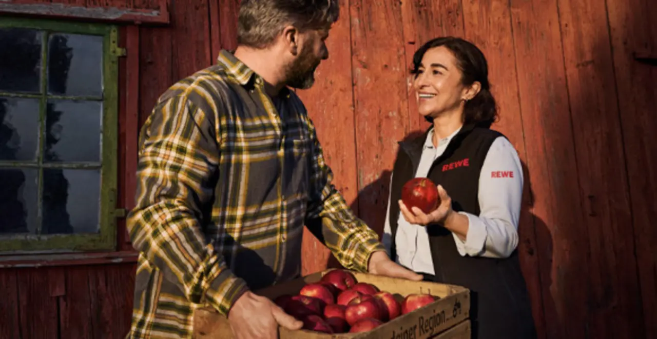 Eine REWE Mitarbeiterin im Gespräch mit einem regionalen Landwirt. Die Darstellung vermittelt: Sie weiß, woher die regionalen Äpfel in ihrem REWE Markt kommen und geht partnerschaftlich mit den Erzeuger:innen um.
