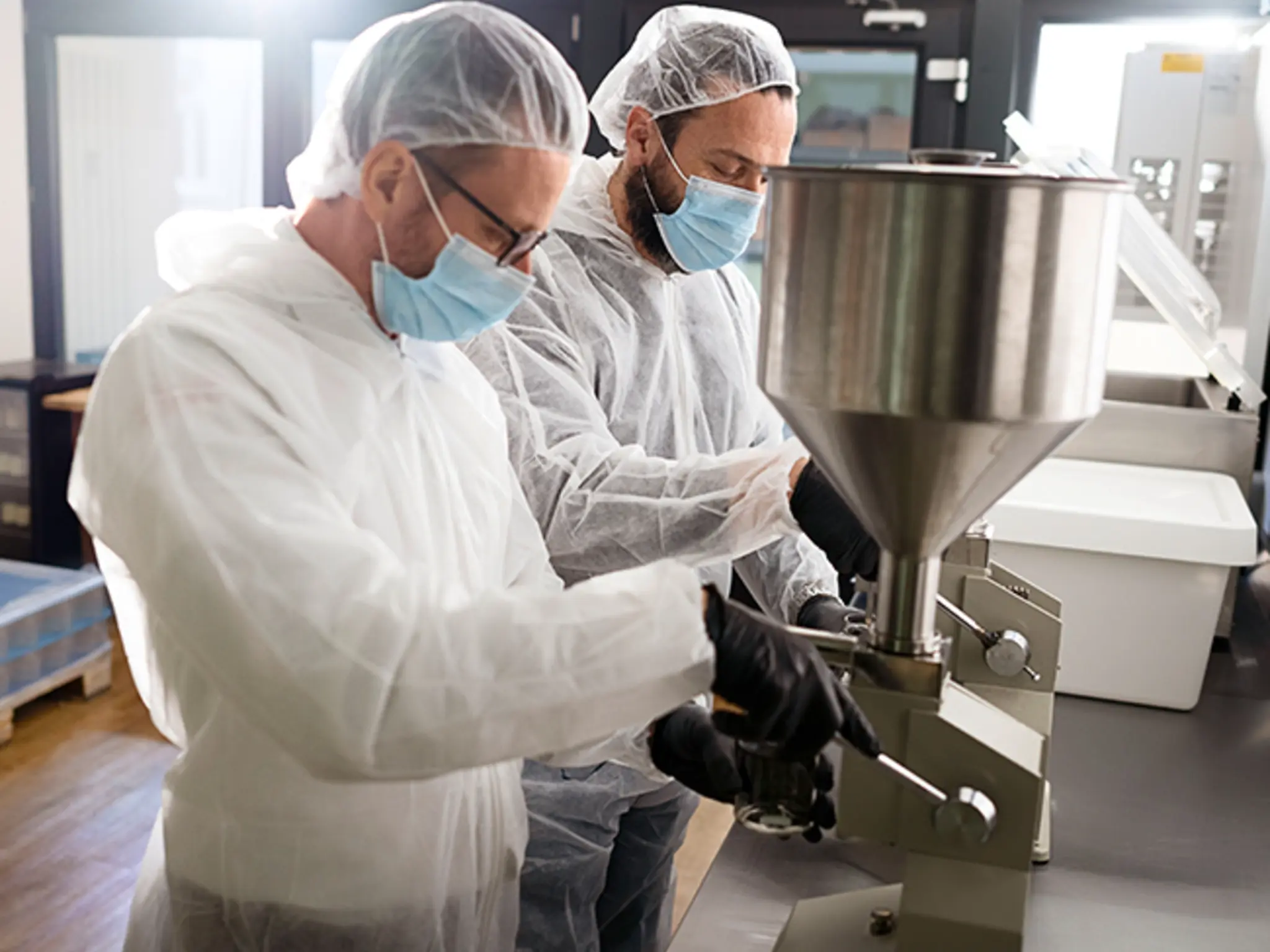 Robert Buljat und Andre Becker mit Mundschutz und Haarnetz in der Großküche. Gemeinsam füllen sie Aioli in Gläser ab.