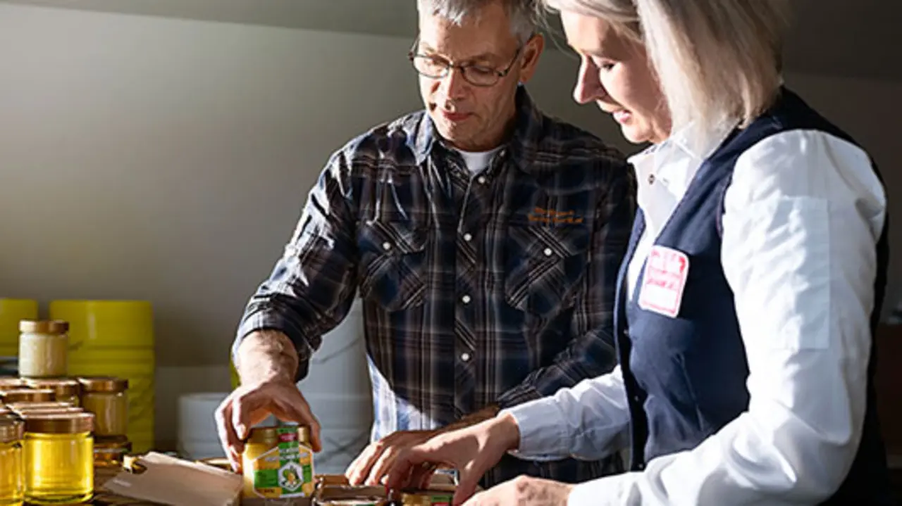 Imker Jan Fischer und REWE Kauffrau Olga Berg verpacken regionale Honiggläser in Pappkartons. 