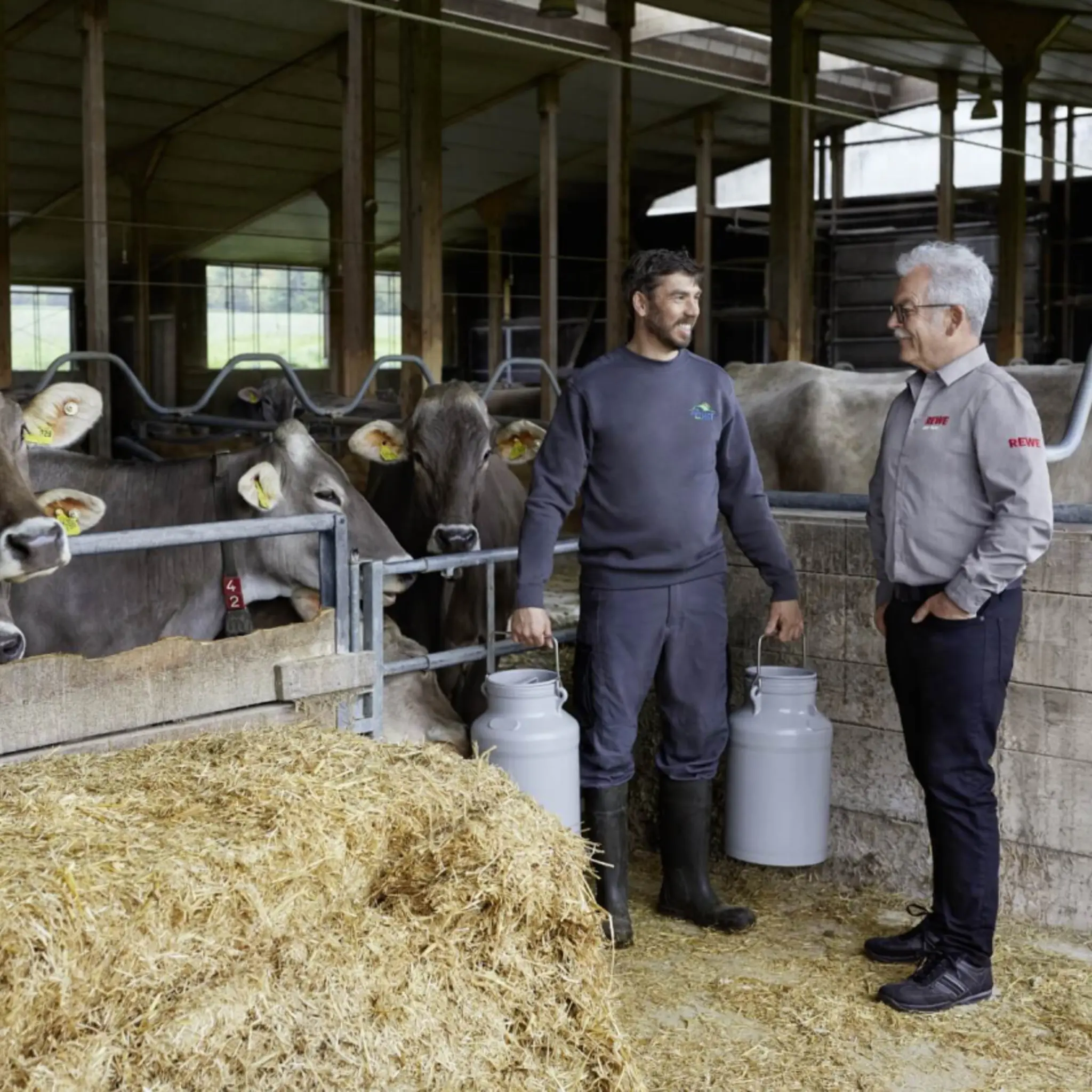 Michael Müller und REWE Kaufmann Rainer Hahn im Kuhstall des Allgäu-Hofs. Michael Müller trägt zwei große Milchkannen.