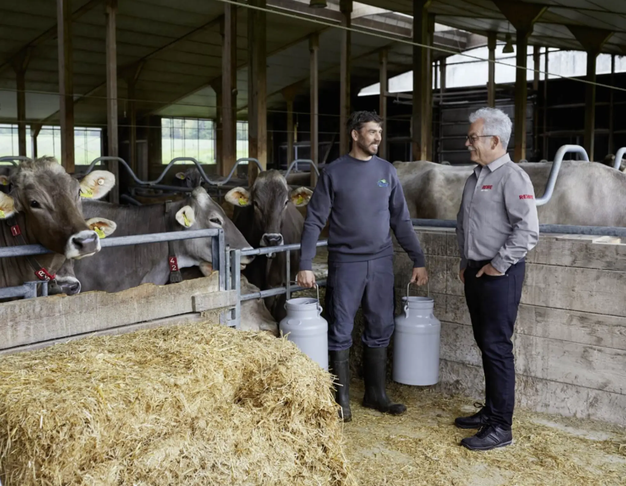 Michael Müller und REWE Kaufmann Rainer Hahn im Kuhstall des Allgäu-Hofs. Michael Müller trägt zwei große Milchkannen.