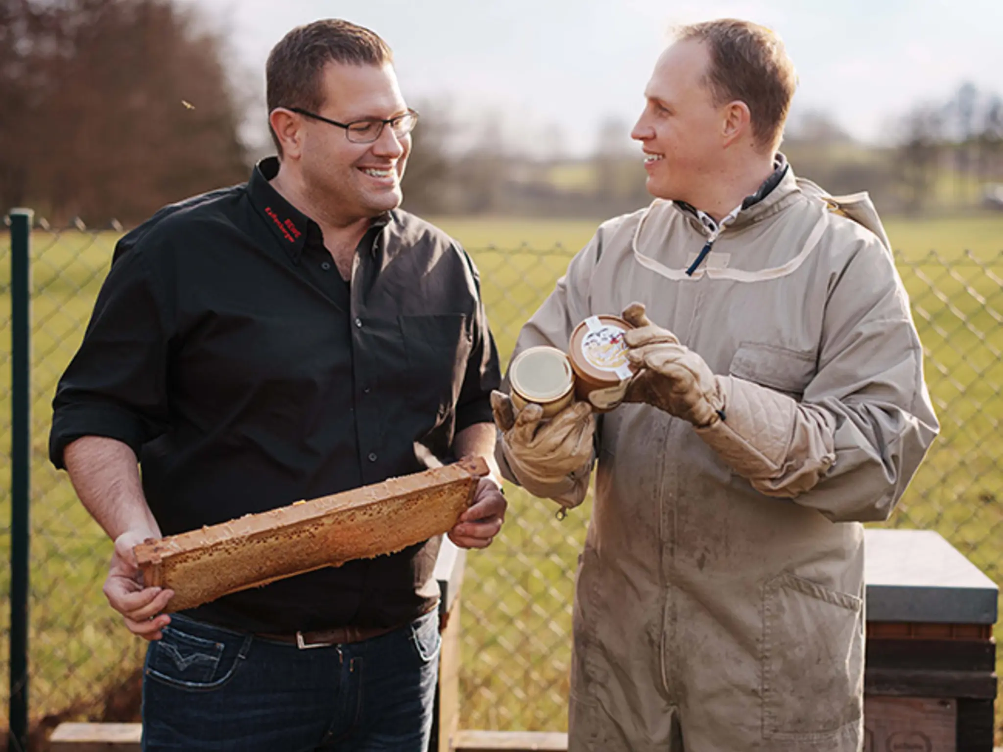 Sascha Greiner und Bernd Kaffenberger stehen auf einer Wiese vor den Bienenstöcken und betrachten eine Honigwabe.
