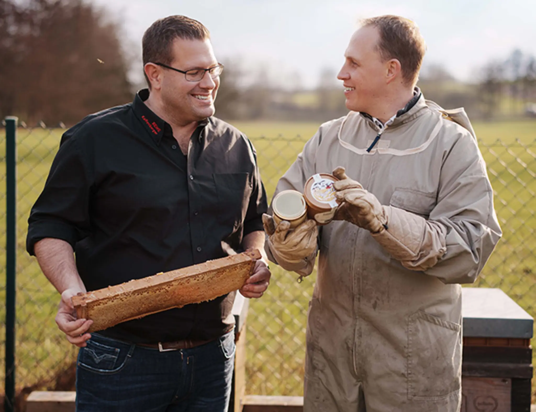 Sascha Greiner und Bernd Kaffenberger stehen auf einer Wiese vor den Bienenstöcken und betrachten eine Honigwabe.
