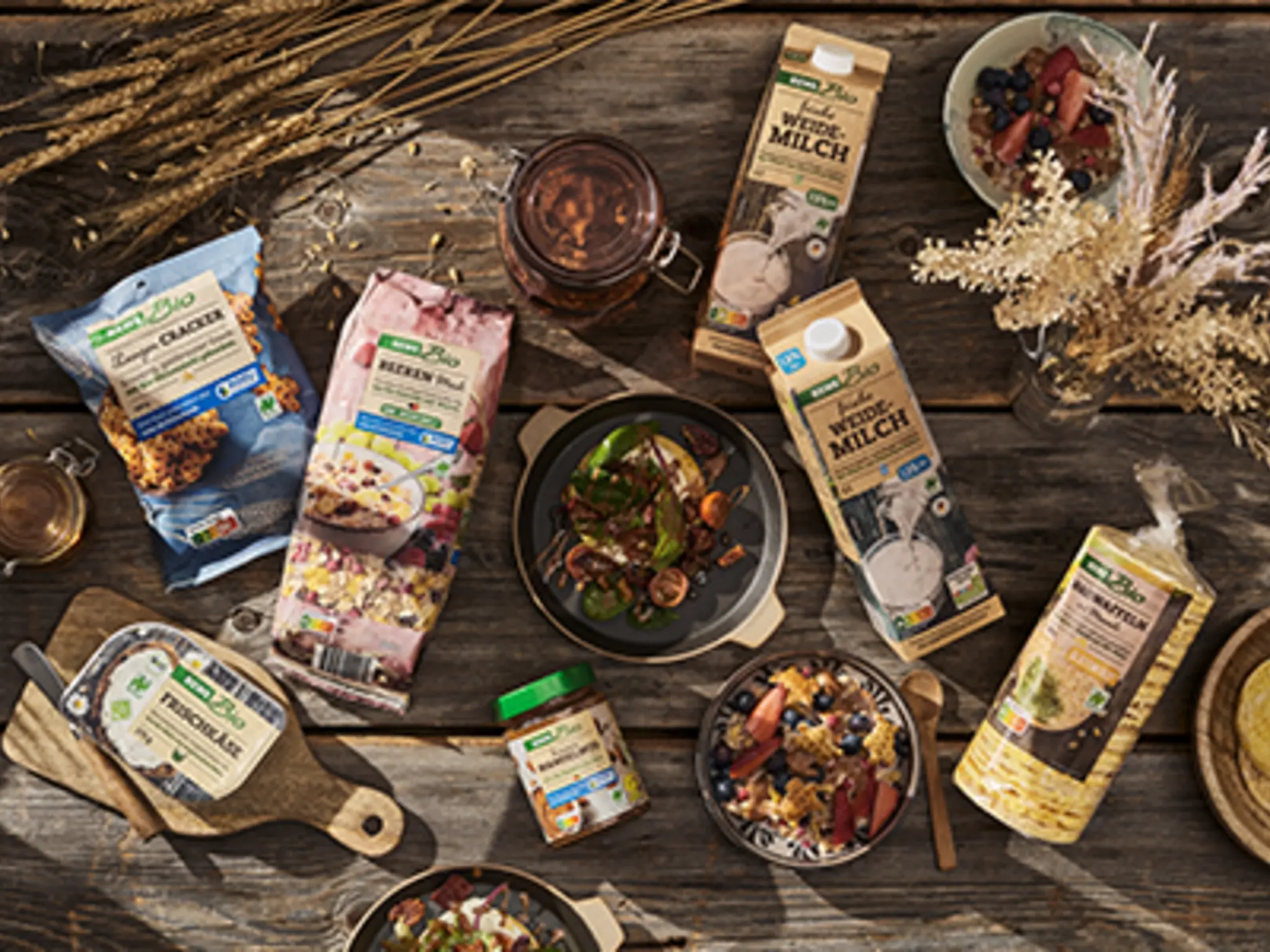 Eine Auswahl an REWE Bio Produkten mit Naturland Zeichen liegen auf einem rustikalen Holztisch. Dazwischen stehen mehrere Schüsseln mit unterschiedlichen Gerichten und reife Kornähren.