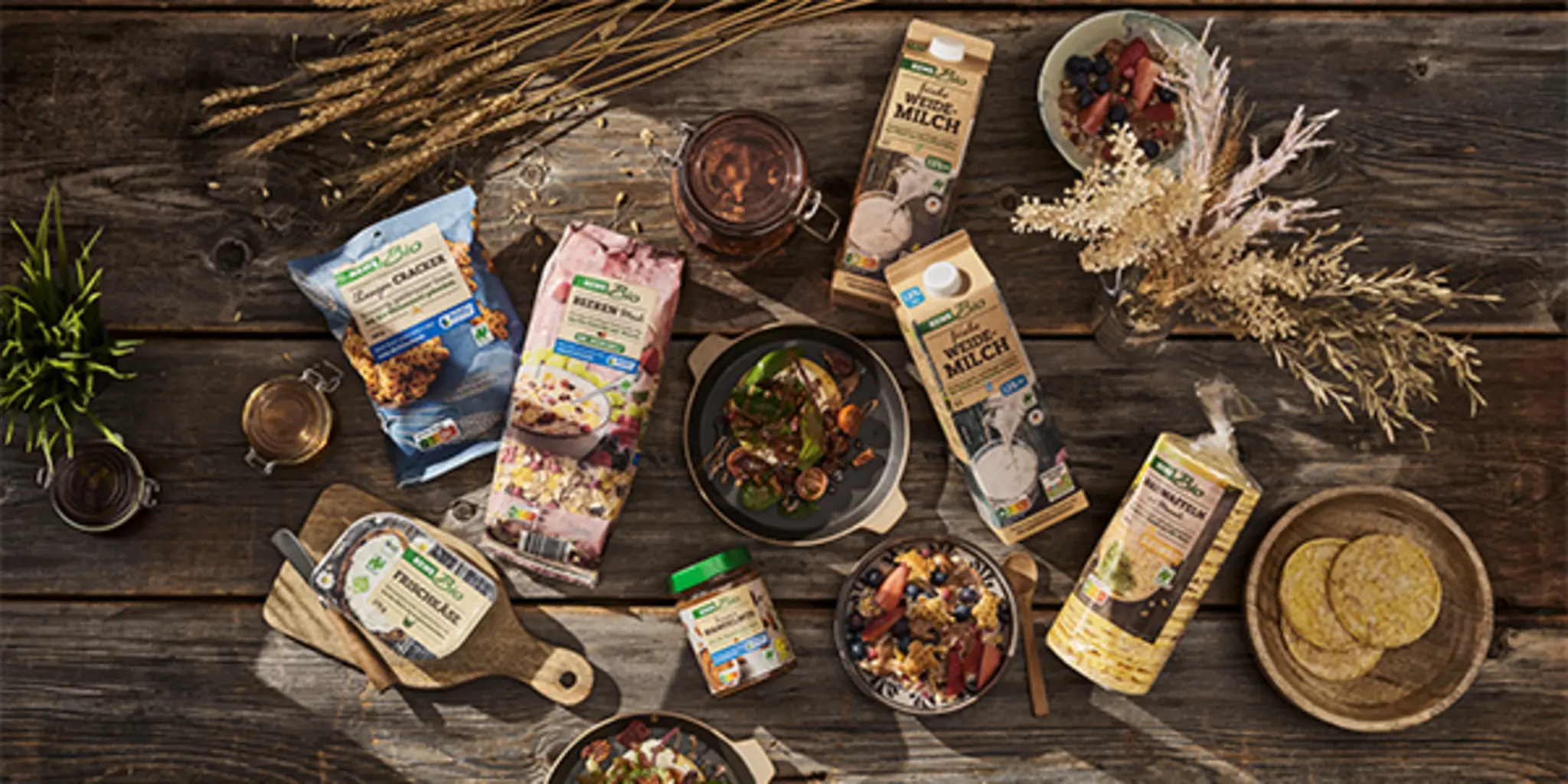 Eine Auswahl an REWE Bio Produkten mit Naturland Zeichen liegen auf einem rustikalen Holztisch. Dazwischen stehen mehrere Schüsseln mit unterschiedlichen Gerichten und reife Kornähren.