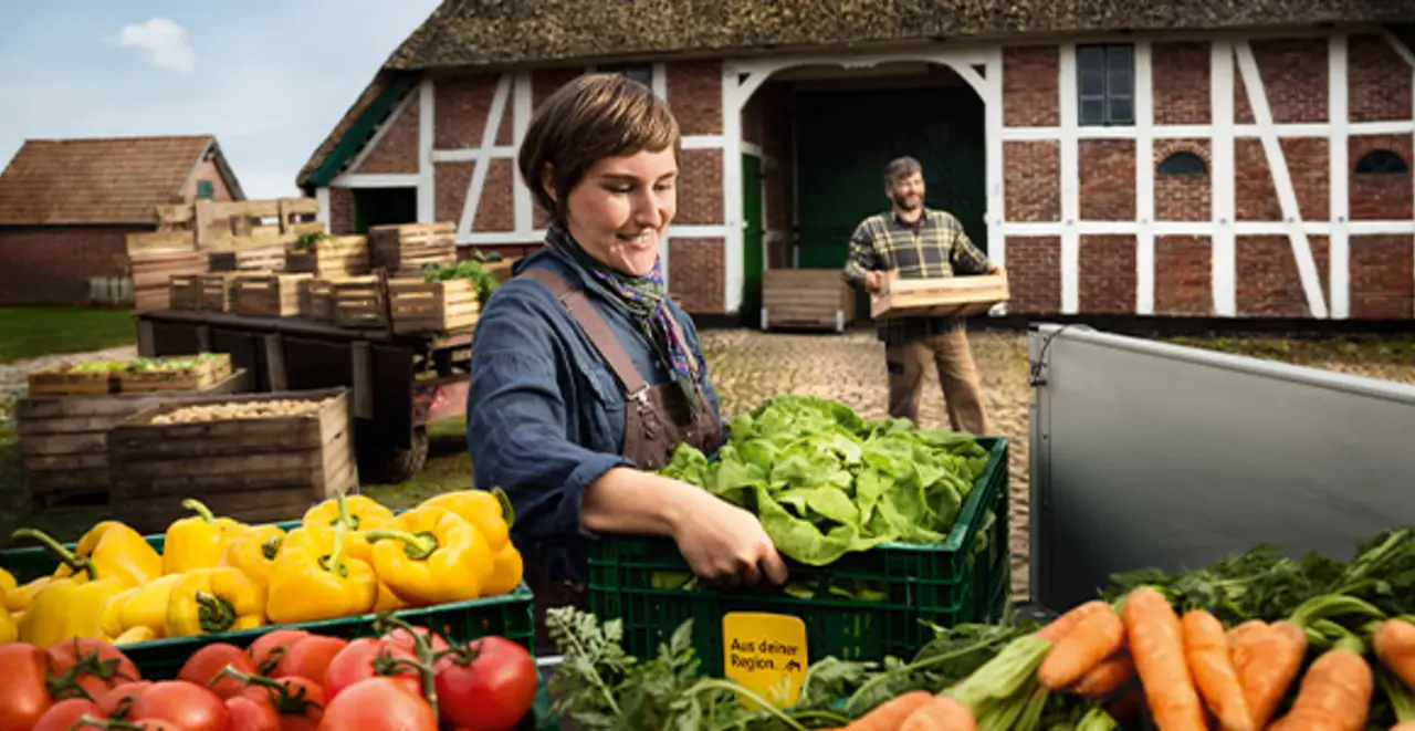 Regionale Erzeuger:innern laden Obst und Gemüse auf einen Anhänger. Zwischen den Produkten steckt das gelbe Schildchen mit dem Schriftzug „Aus deiner Region“, das du auch im Markt findest. 