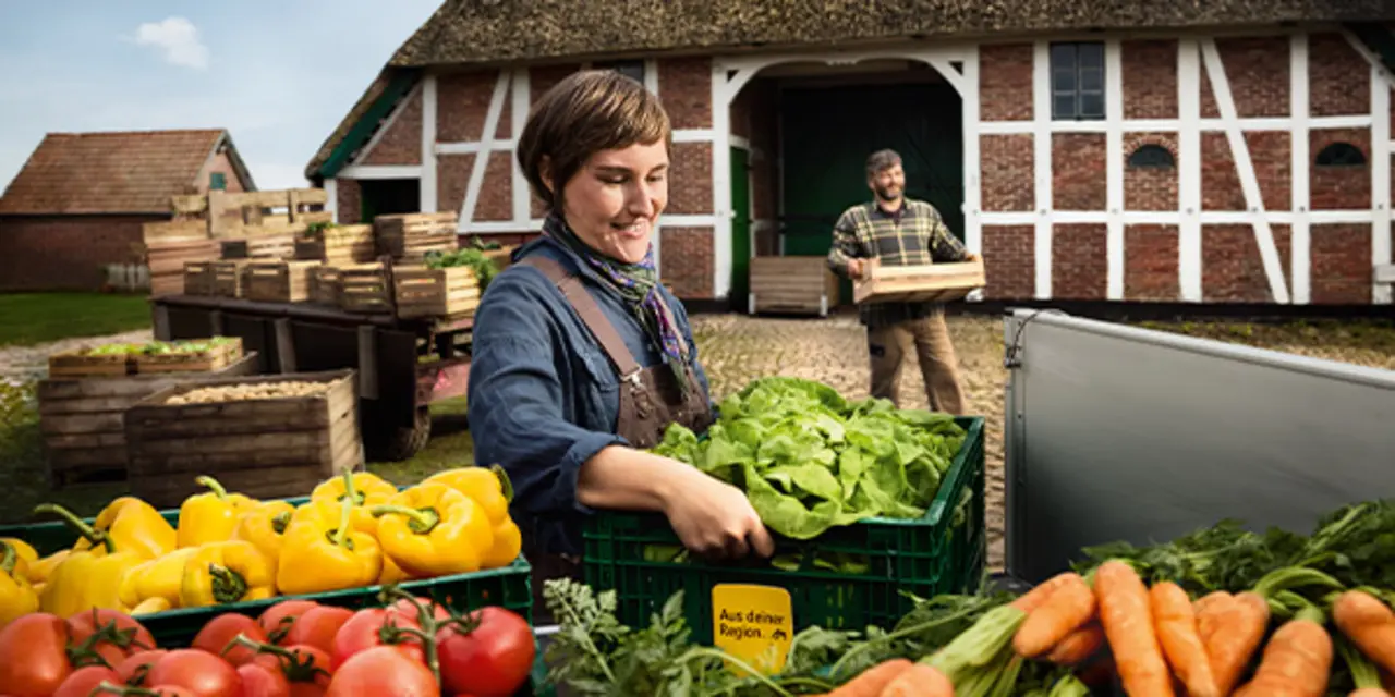 Regionale Erzeuger:innern laden Obst und Gemüse auf einen Anhänger. Zwischen den Produkten steckt das gelbe Schildchen mit dem Schriftzug „Aus deiner Region“, das du auch im Markt findest.