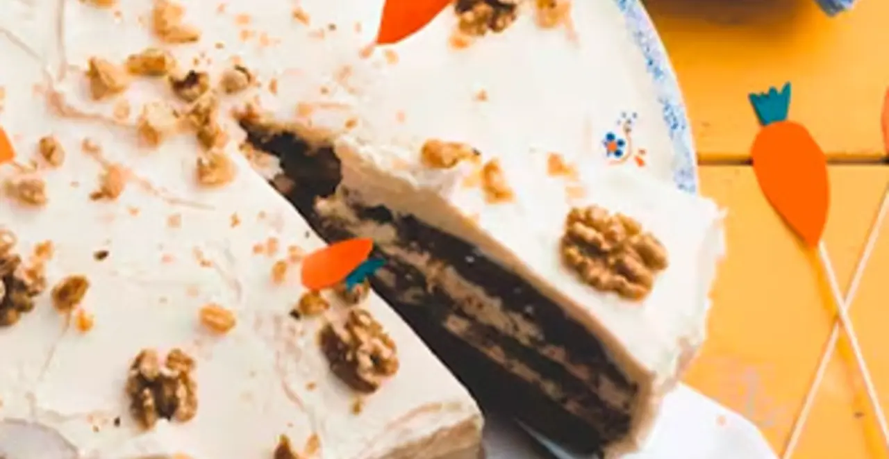 Ein Carrot Cake ohne Eier steht auf einem Tisch. Jemand dekoriert ihn gerade mit Karotten aus Pappe. Ein Stück Kuchen fehlt schon. 