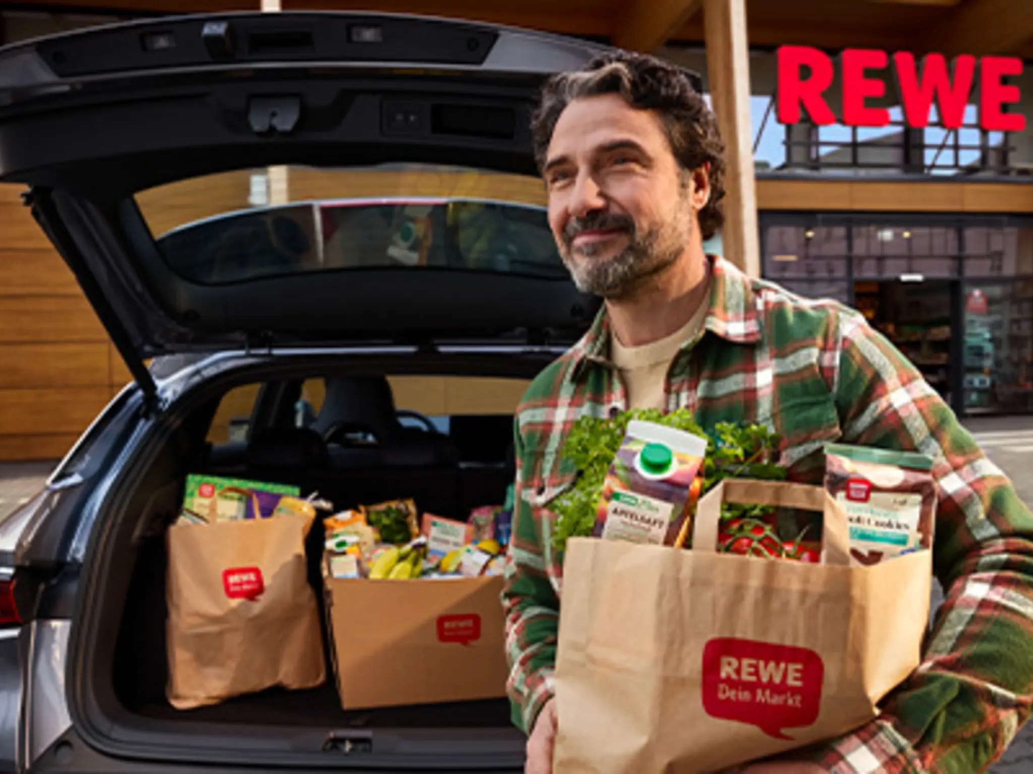 Auf dem Parkplatz vor einem REWE Markt: Eine Person lädt volle Einkaufstüten in den Kofferraum.
