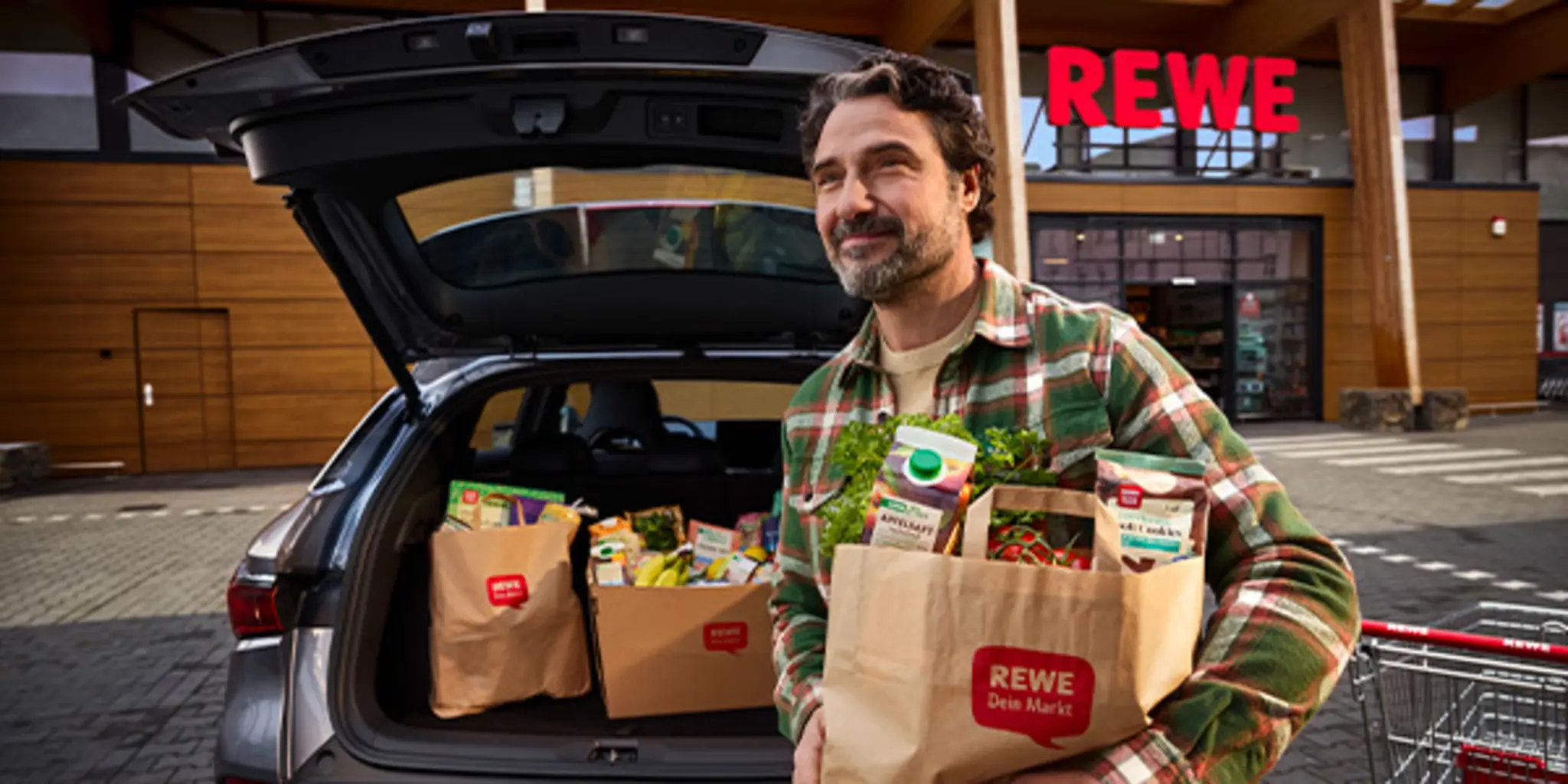 Auf dem Parkplatz vor einem REWE Markt: Eine Person lädt volle Einkaufstüten in den Kofferraum. 