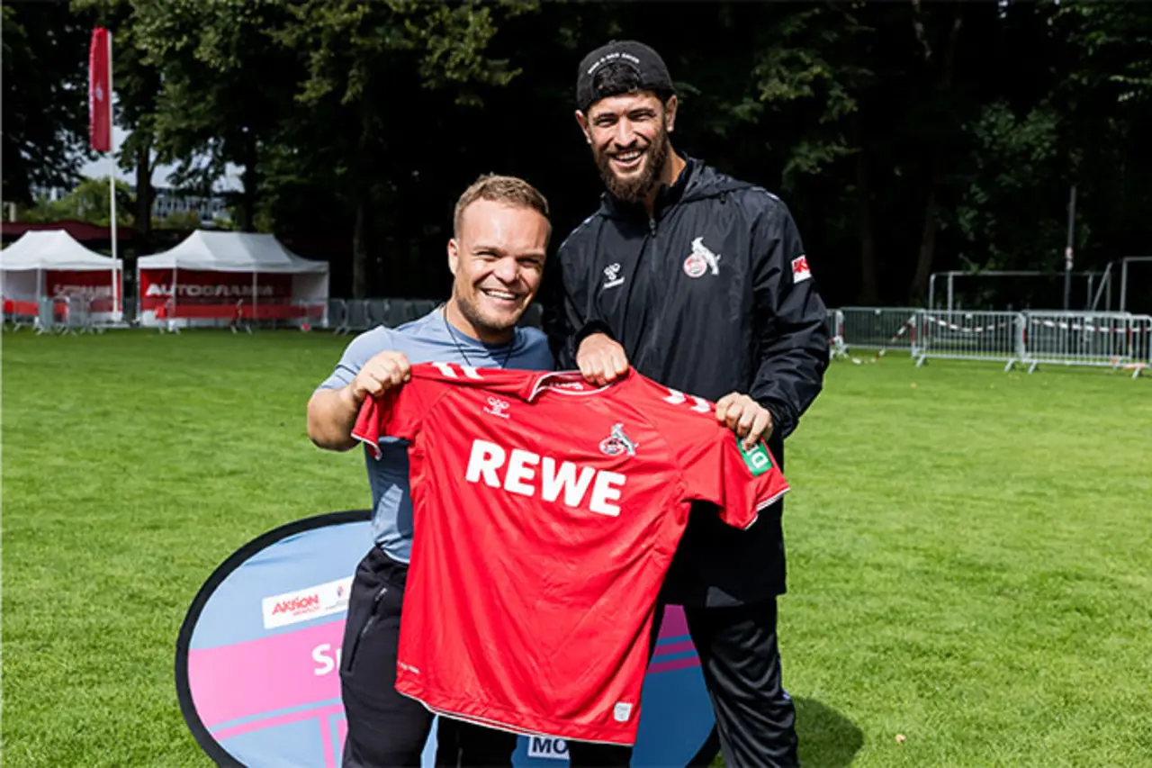 Zwei Männer stehen auf einem Fußballplatz und halten gemeinsam ein rotes FC-Trikot mit der Aufschrift ‚REWE‘ in die Kamera; im Hintergrund sind Absperrungen und Veranstaltungsbanner zu sehen. Copyright: Steffie Wunderlich