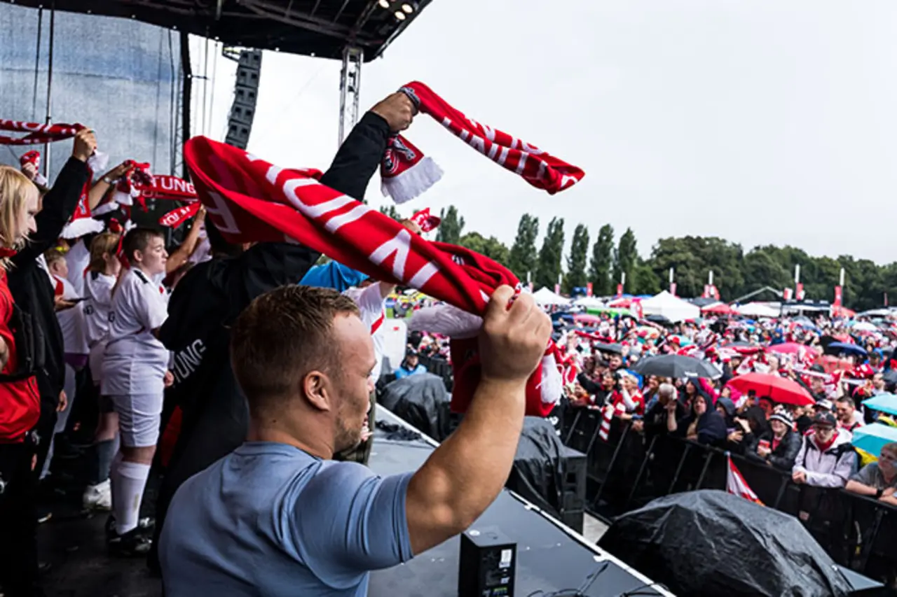 Personen auf einer Bühne schwenken rote Fanschals in Richtung einer großen Menschenmenge vor der Bühne; viele Zuschauer stehen mit Regenschirmen auf einem Veranstaltungsgelände im Freien. Copyright: Steffie Wunderlich
