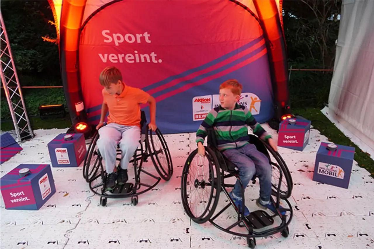Zwei Jungen sitzen in Sportrollstühlen vor einem beleuchteten Banner mit der Aufschrift ‚Sport vereint.‘ und bewegen sich auf einer Aktionsfläche bei einer inklusiven Veranstaltung.
