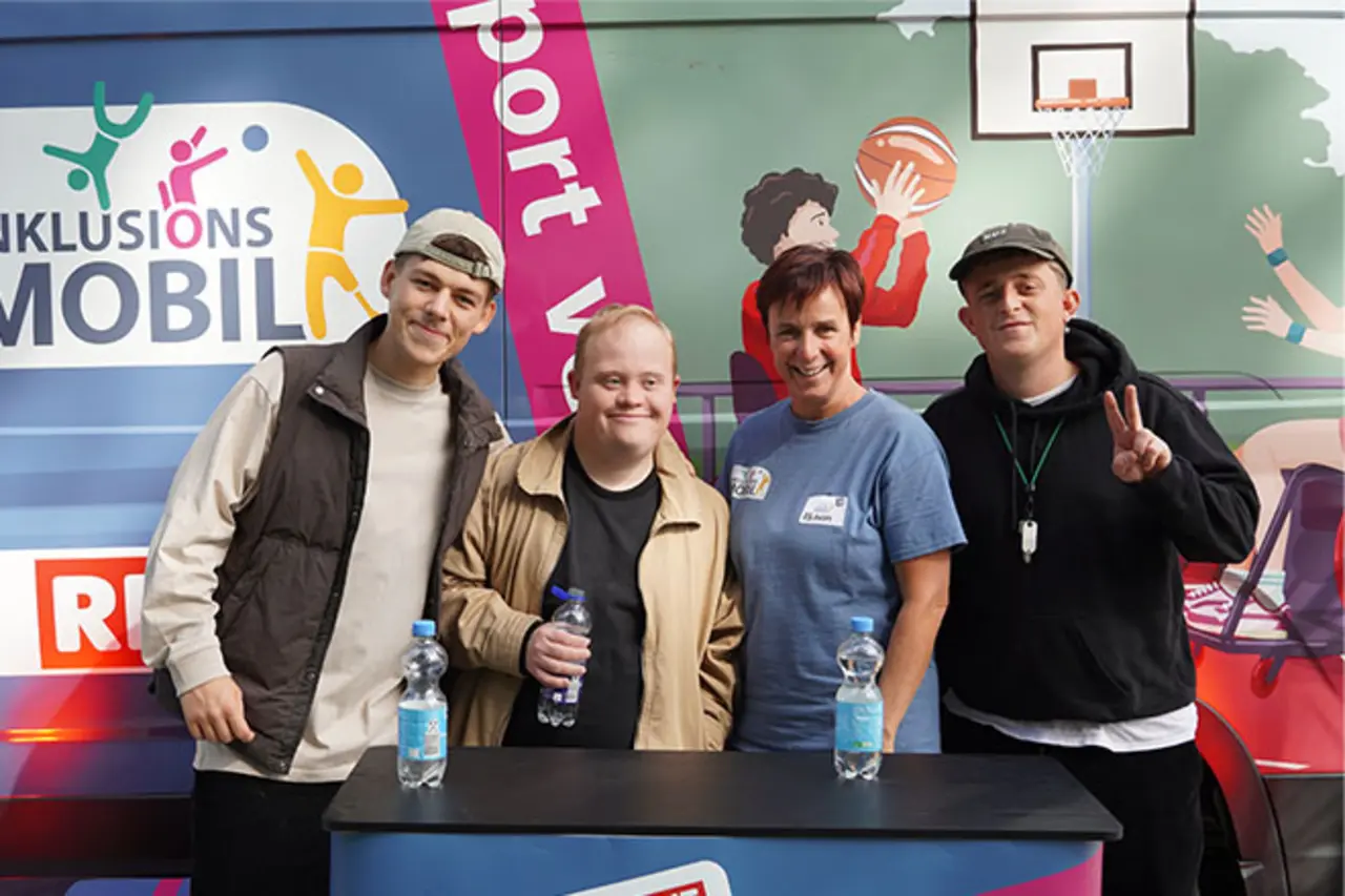 Vier Personen stehen vor einem Informationsstand und einem Fahrzeug mit der Aufschrift ‚Inklusionsmobil – Sport vereint.‘; sie lächeln in die Kamera, zwei halten Wasserflaschen in der Hand. Copyright: Achim Bachhausen