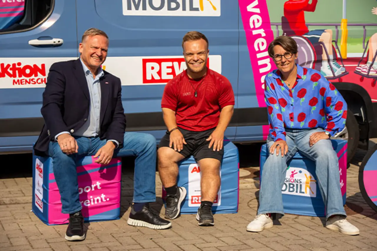 Drei Personen sitzen auf Sitzwürfeln vor einem gebrandeten Fahrzeug mit der Aufschrift ‚Inklusionsmobil‘; im Hintergrund sind Logos wie ‚REWE‘ und ein Banner mit dem Slogan ‚Sport vereint.‘ zu sehen. Copyright: Steffie Wunderlich