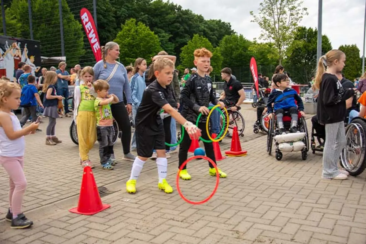 Kinder nehmen an einer Mitmachstation im Freien teil und werfen bunte Ringe zwischen Hütchen; im Hintergrund sind weitere Kinder, Erwachsene und Rollstuhlnutzende bei einer inklusiven Sportveranstaltung zu sehen. Copyright: Achim Bachhausen