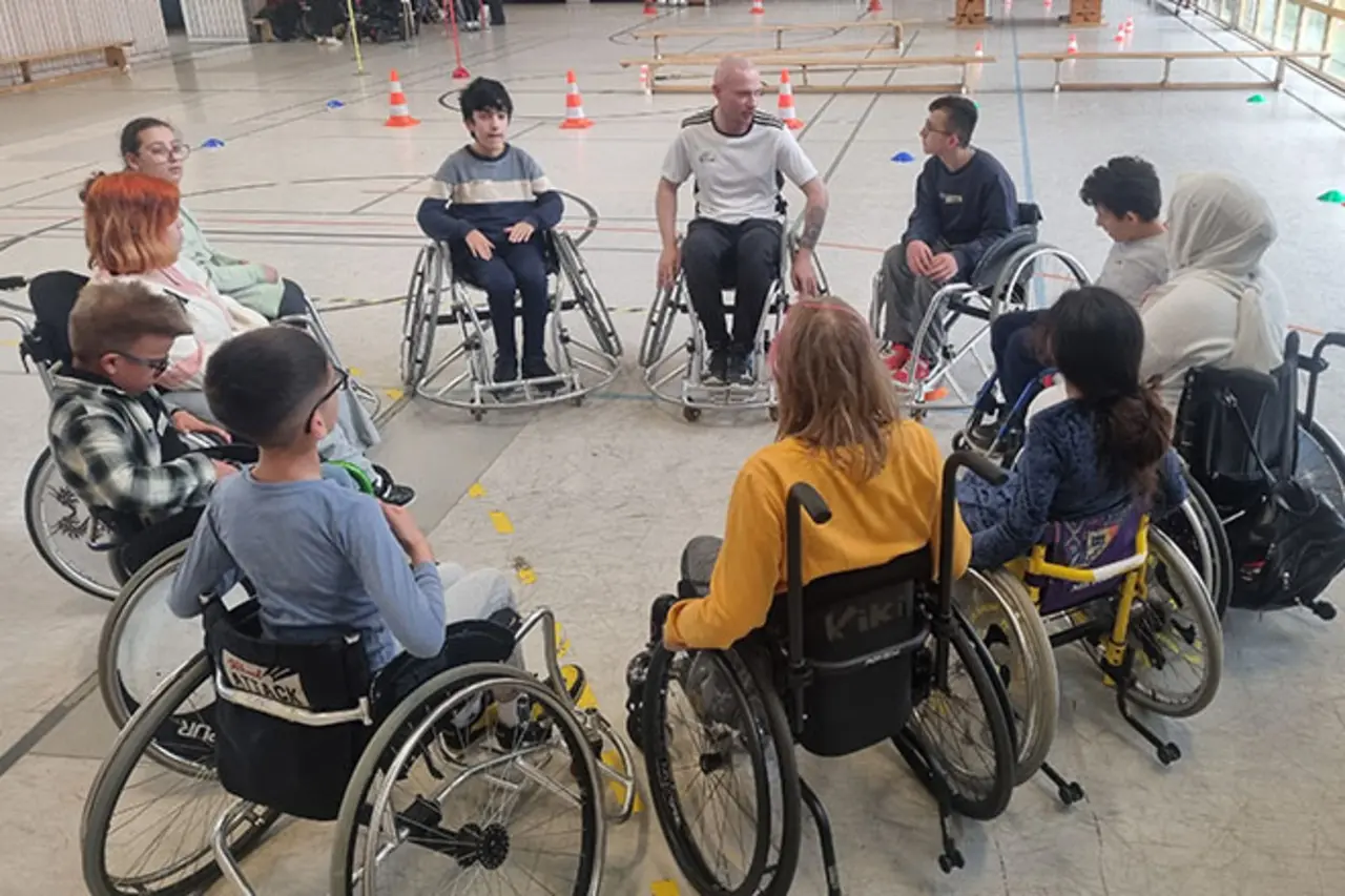 Gruppe von Kindern und einem Trainer im Rollstuhl sitzen im Kreis in einer Sporthalle mit Parcours-Hütchen und Trainingsaufbau Copyright:DBS
