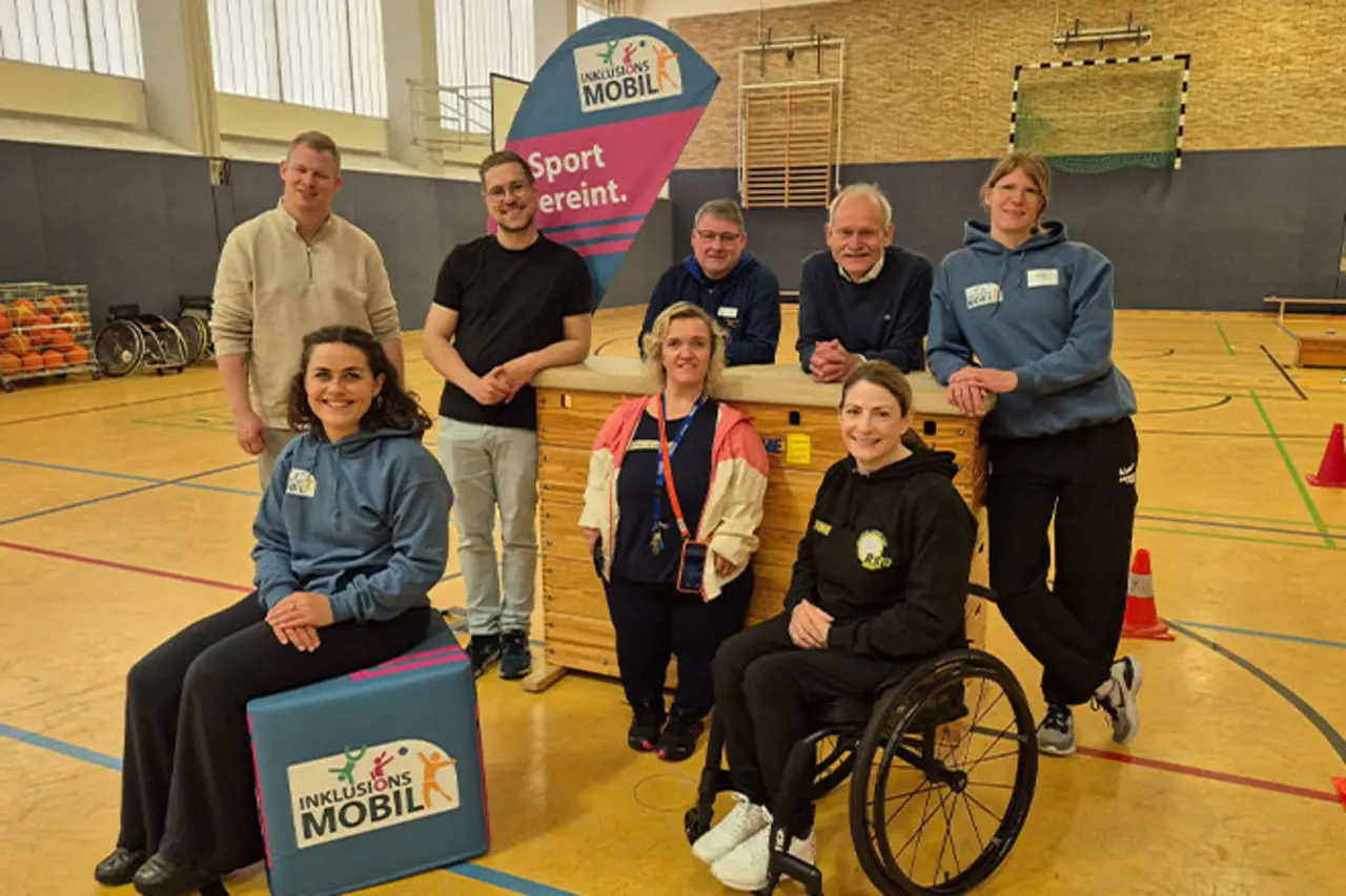 Acht Erwachsene, darunter eine Frau im Rollstuhl, posieren in einer Sporthalle vor einem Banner mit der Aufschrift ‚Inklusionsmobil – Sport vereint.‘ und einer Turngeräte-Box. Copyright: DBS