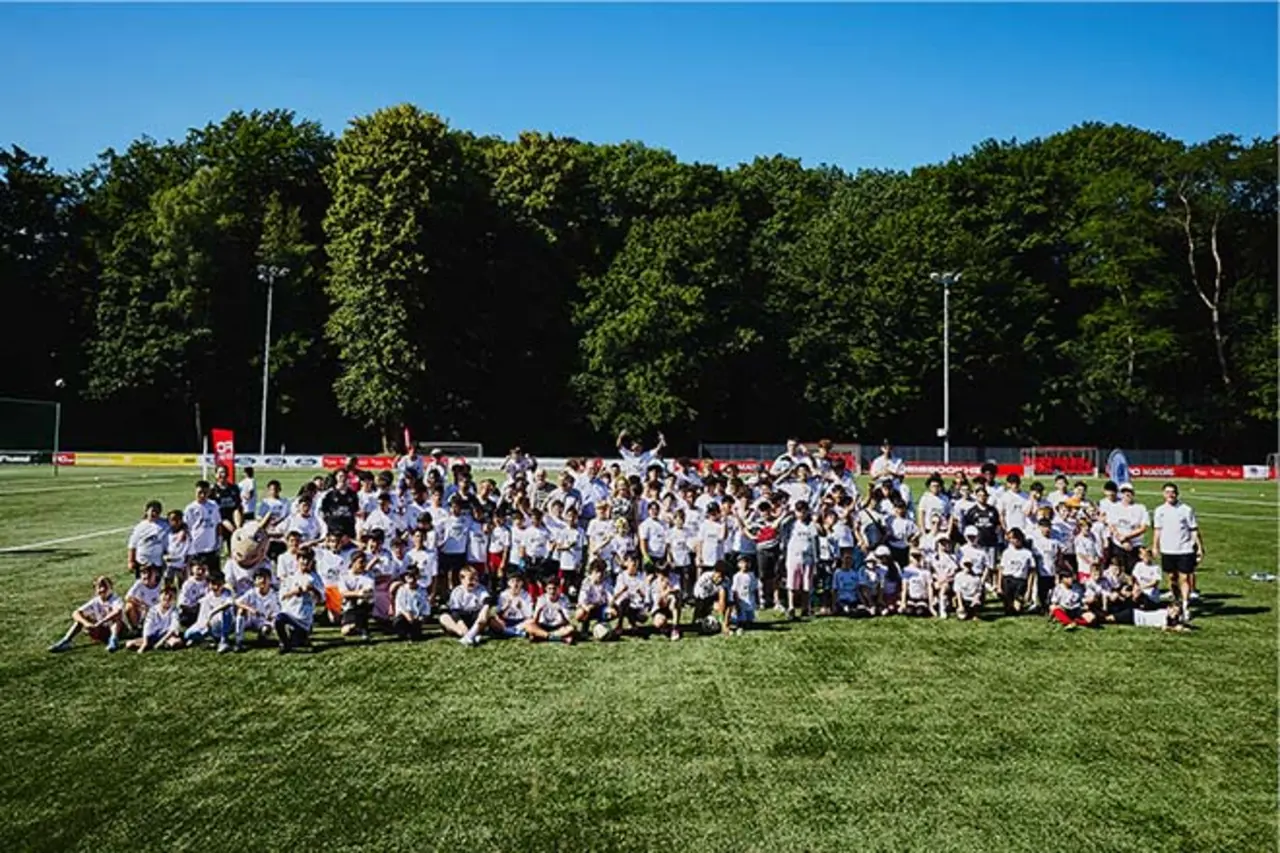 Große Gruppe von Kindern und Erwachsenen in weißen T-Shirts posiert gemeinsam auf einem Sportplatz vor einer Baumreihe bei sonnigem Wetter. Copyright: DBS