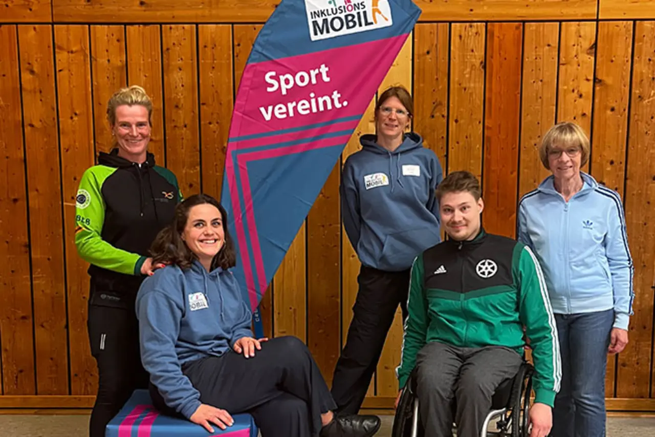 Fünf Erwachsene, darunter ein Mann im Rollstuhl, stehen und sitzen vor einer Holzwand neben einem Banner mit der Aufschrift ‚Inklusionsmobil – Sport vereint.