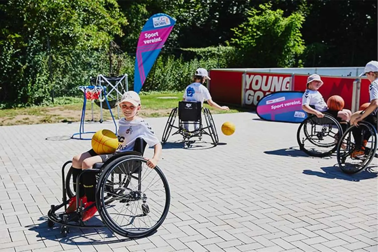 Kinder im Rollstuhl spielen Basketball auf einem gepflasterten Platz im Freien; im Hintergrund stehen Banner mit der Aufschrift ‚Inklusionsmobil – Sport vereint. Copyright: Jannik Czauderna