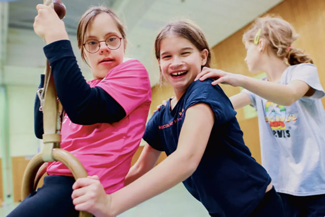 Ein Kind mit Behinderung schwingt in einer Turnhalle an einem Tau. Ein zweites Kind hilft ihm. Copyright: TV-Ratingen (Alt-Tag 2) Gruppe von Kindern und einem Trainer im Rollstuhl sitzen im Kreis in einer Sporthalle mit Parcours-Hütchen und Trainingsaufbau Copyright:DBS (Alt-Tag 3) Fünf Erwachsene, darunter ein Mann im Rollstuhl, stehen und sitzen vor einer Holzwand neben einem Banner mit der Aufschrift ‚Inklusionsmobil – Sport vereint. (Alt-Tag 4) Sporthalle mit aufgebautem Rollstuhl-Parcours, Hütchen und Helmen auf einem Tisch; im Vordergrund ein Banner mit der Aufschrift ‚Inklusionsmobil – Sport vereint. Copyright:DBS (Alt-Tag 5) Acht Erwachsene, darunter eine Frau im Rollstuhl, posieren in einer Sporthalle vor einem Banner mit der Aufschrift ‚Inklusionsmobil – Sport vereint.‘ und einer Turngeräte-Box. Copyright: DBS (Alt-Tag 6) Große Gruppe von Kindern und Erwachsenen in weißen T-Shirts posiert gemeinsam auf einem Sportplatz vor einer Baumreihe bei sonnigem Wetter. Copyright: DBS (Alt-Tag 7) Kinder im Rollstuhl spielen Basketball auf einem gepflasterten Platz im Freien; im Hintergrund stehen Banner mit der Aufschrift ‚Inklusionsmobil – Sport vereint. Copyright: Jannik Czauderna (Alt-Tag 8) Gruppe von Fußballspielern in rot-weißen Trikots und Begleitpersonen posiert gemeinsam auf einem Fußballplatz vor einem Tor bei sonnigem Wetter. Copyright: Jannik Czauderna (Alt-Tag 9) Zwei Männer stehen auf einem Fußballplatz und halten gemeinsam ein rotes FC-Trikot mit der Aufschrift ‚REWE‘ in die Kamera; im Hintergrund sind Absperrungen und Veranstaltungsbanner zu sehen. Copyright: Steffie Wunderlich (Alt-Tag 10) Personen auf einer Bühne schwenken rote Fanschals in Richtung einer großen Menschenmenge vor der Bühne; viele Zuschauer stehen mit Regenschirmen auf einem Veranstaltungsgelände im Freien. Copyright: Steffie Wunderlich (Alt-Tag 11) Drei Personen sitzen auf Sitzwürfeln vor einem gebrandeten Fahrzeug mit der Aufschrift ‚Inklusionsmobil‘; im Hintergrund sind Logos wie ‚REWE‘ und ein Banner mit dem Slogan ‚Sport vereint.‘ zu sehen. Copyright: Steffie Wunderlich (Alt-Tag 12) Kinder nehmen an einer Mitmachstation im Freien teil und werfen bunte Ringe zwischen Hütchen; im Hintergrund sind weitere Kinder, Erwachsene und Rollstuhlnutzende bei einer inklusiven Sportveranstaltung zu sehen. Copyright: Achim Bachhausen (Alt-Tag 13) Vier Personen stehen vor einem Informationsstand und einem Fahrzeug mit der Aufschrift ‚Inklusionsmobil – Sport vereint.‘; sie lächeln in die Kamera, zwei halten Wasserflaschen in der Hand. Copyright: Achim Bachhausen (Alt-Tag 14) Gruppe von Erwachsenen, darunter zwei Personen im Rollstuhl, posiert gemeinsam auf einer Bühne beim Bürgerfest. Copyright: DBS (Alt-Tag 15) Zwei Jungen sitzen in Sportrollstühlen vor einem beleuchteten Banner mit der Aufschrift ‚Sport vereint.‘ und bewegen sich auf einer Aktionsfläche bei einer inklusiven Veranstaltung.