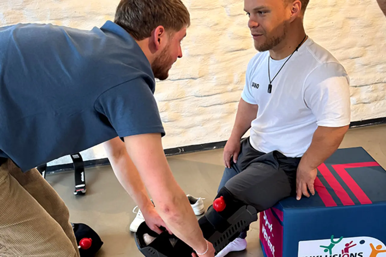 Para-Kugelstoßer Niko Kappel sitzt auf einem niedrigen Hocker. Ein Helfer legt ihm eine Beinorthese an.