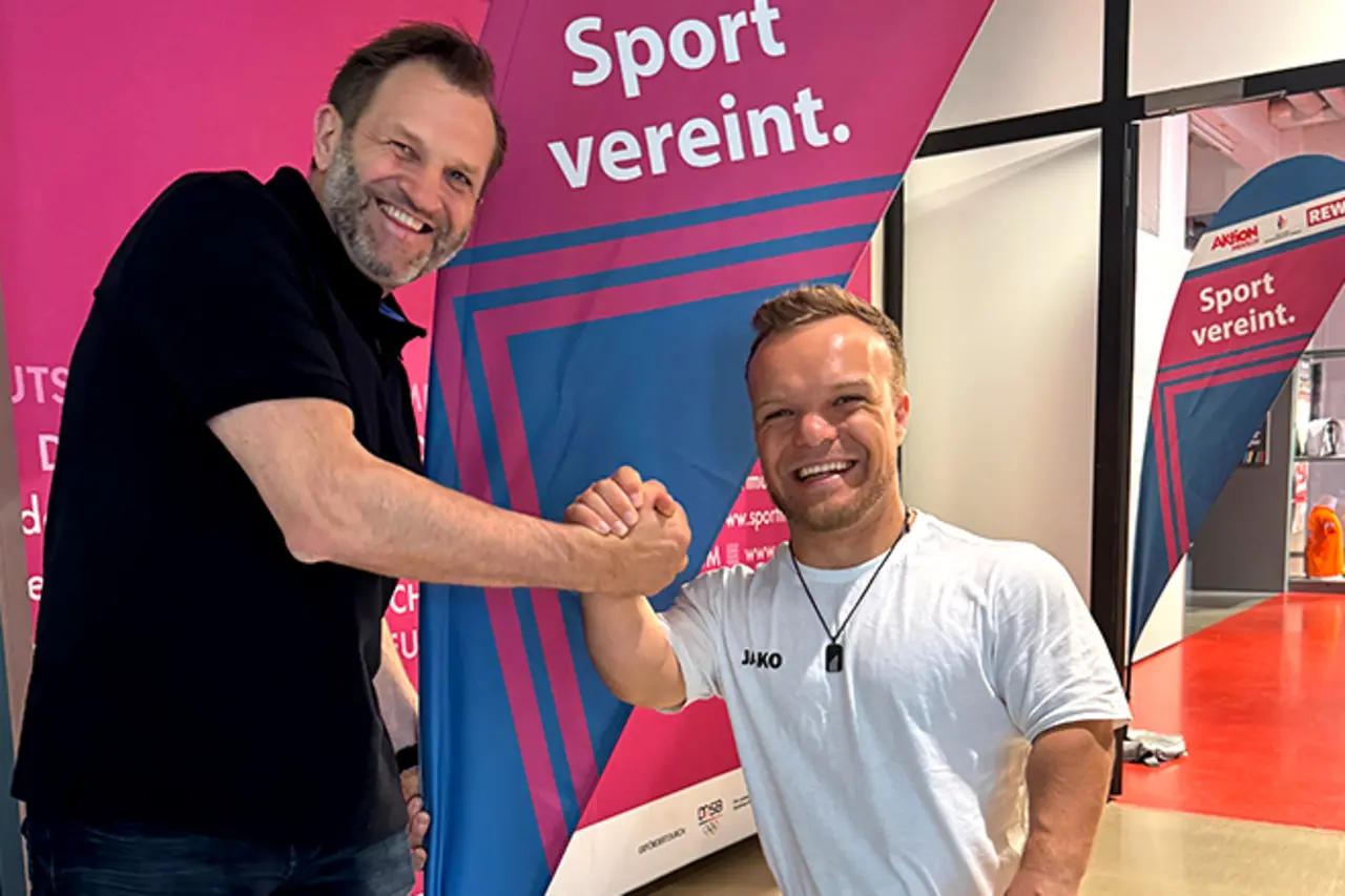Para-Kugelstoßer Niko Kappel und ein Verantwortlicher der Veranstaltung beim Handschlag. Auf einer Fahne im Hintergrund steht der Schriftzug „Sport vereint.“