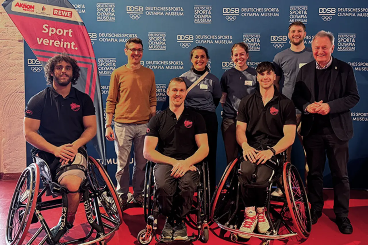 Mehrere Teilnehmende mit und ohne Behinderung vor einer Logo-Wand des Deutschen Sport & Olympia Museum. Auf einer Fahne neben ihnen steht der Schriftzug „Sport vereint“.