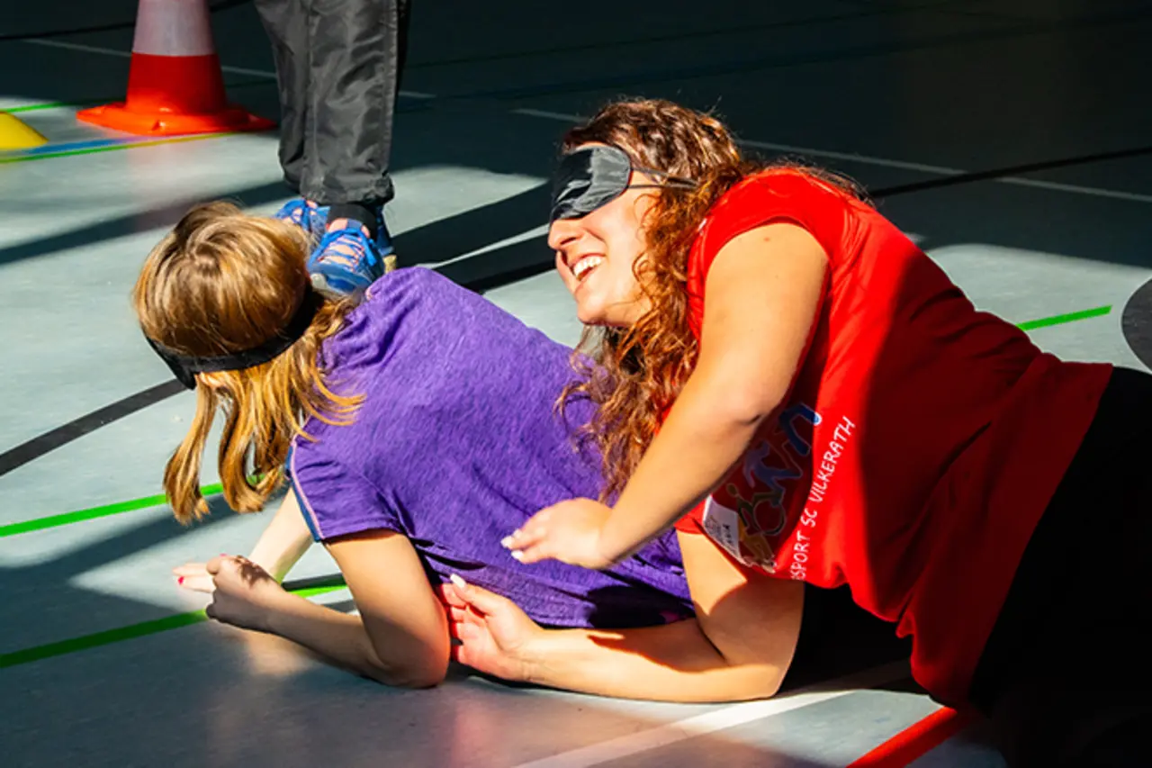 Beim Besuch des Inklusionsmobils trainieren Jugendliche mit Augenbinden. Zwei Mädchen liegen lachend auf dem Boden einer Turnhalle.