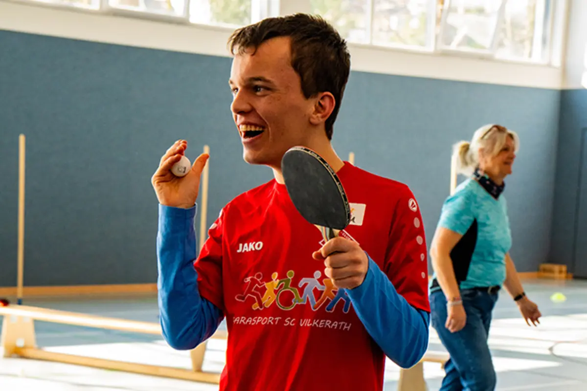 Ein Jugendlicher mit Behinderung trägt ein T-Shirt mit der Aufschrift „Parasport SC Vilkerath“. Er spielt in der Turnhalle Tischtennis. 