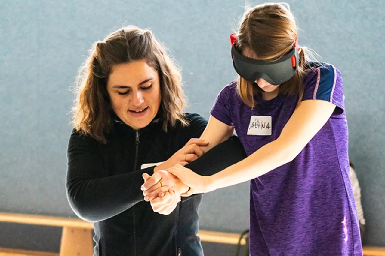 Beim Besuch des Inklusionsmobils in der Turnhalle des SC Vilkerath. Eine Jugendliche mit VR-Brille scheint zu balancieren. Eine Trainerin hält sie am Arm und unterstützt sie.