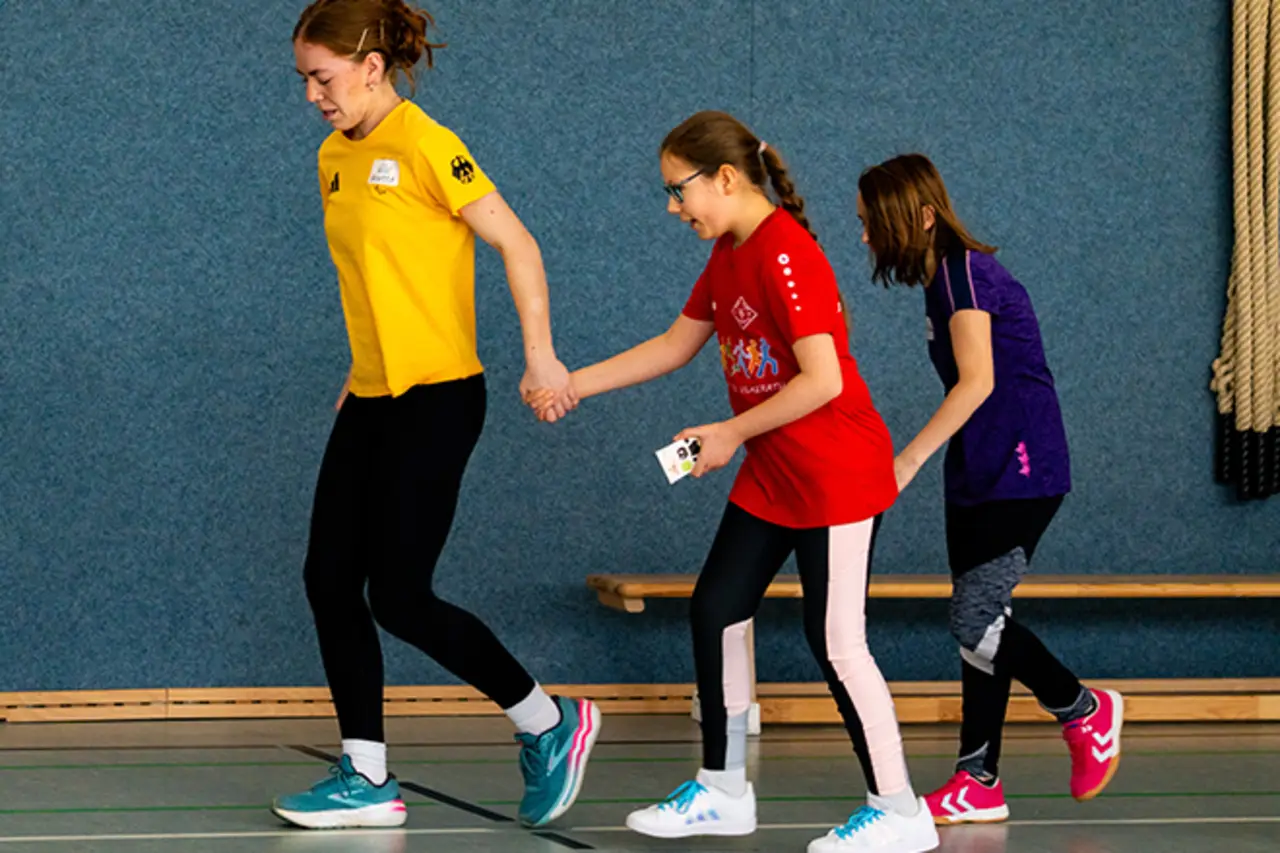 Eine Helferin führt eine Jugendliche mit Behinderung an der Hand durch die Turnhalle. Ein zweites Mädchen folgt den beiden.