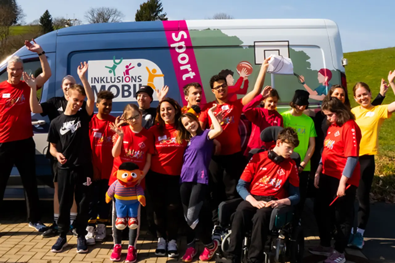 Das Inklusionsmobil auf dem Parkplatz des SC Vilkerath. Viele Teilnehmende mit und ohne Behinderung haben sich versammelt und winken fröhlich.