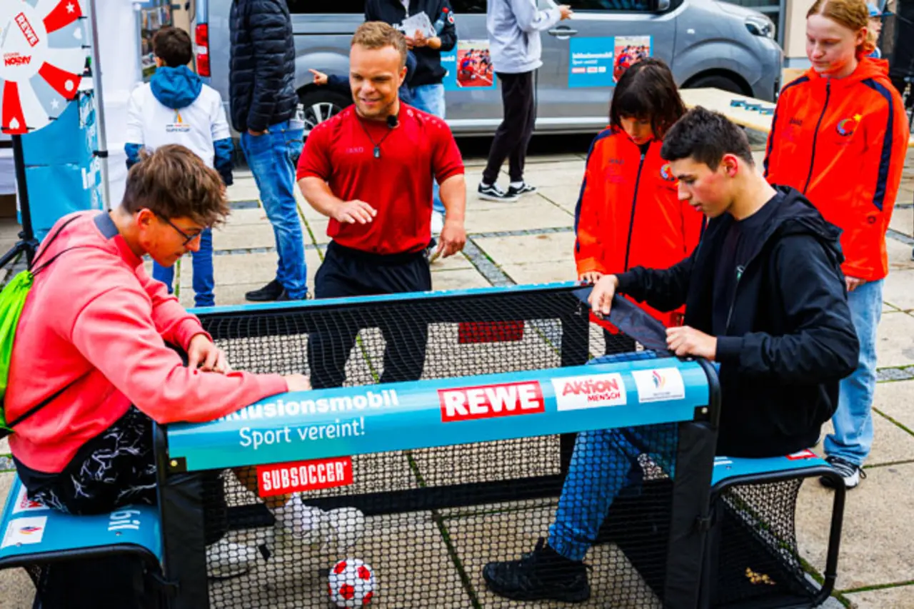 Mehrere Jugendliche mit und ohne Behinderung spielen an einem inklusiven Sportgerät Fußball. Inklusionsmobil Botschafter Niko Kappel gibt Tipps.