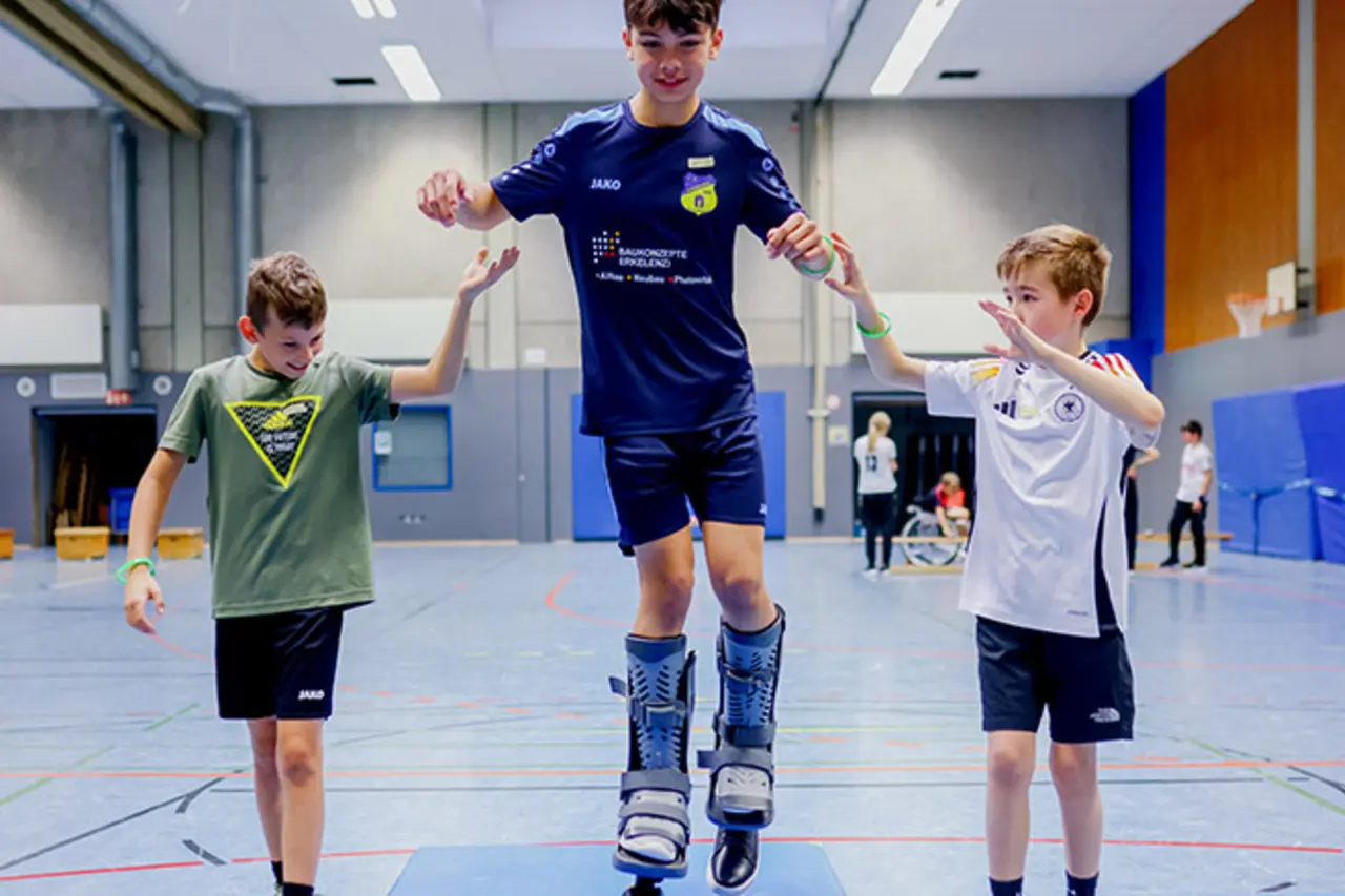 Ein Kind trägt Beinprothesen und balanciert damit auf dem Oberteil eines Turnkastens. Zwei andere Kinder helfen beim Gleichgewicht halten.