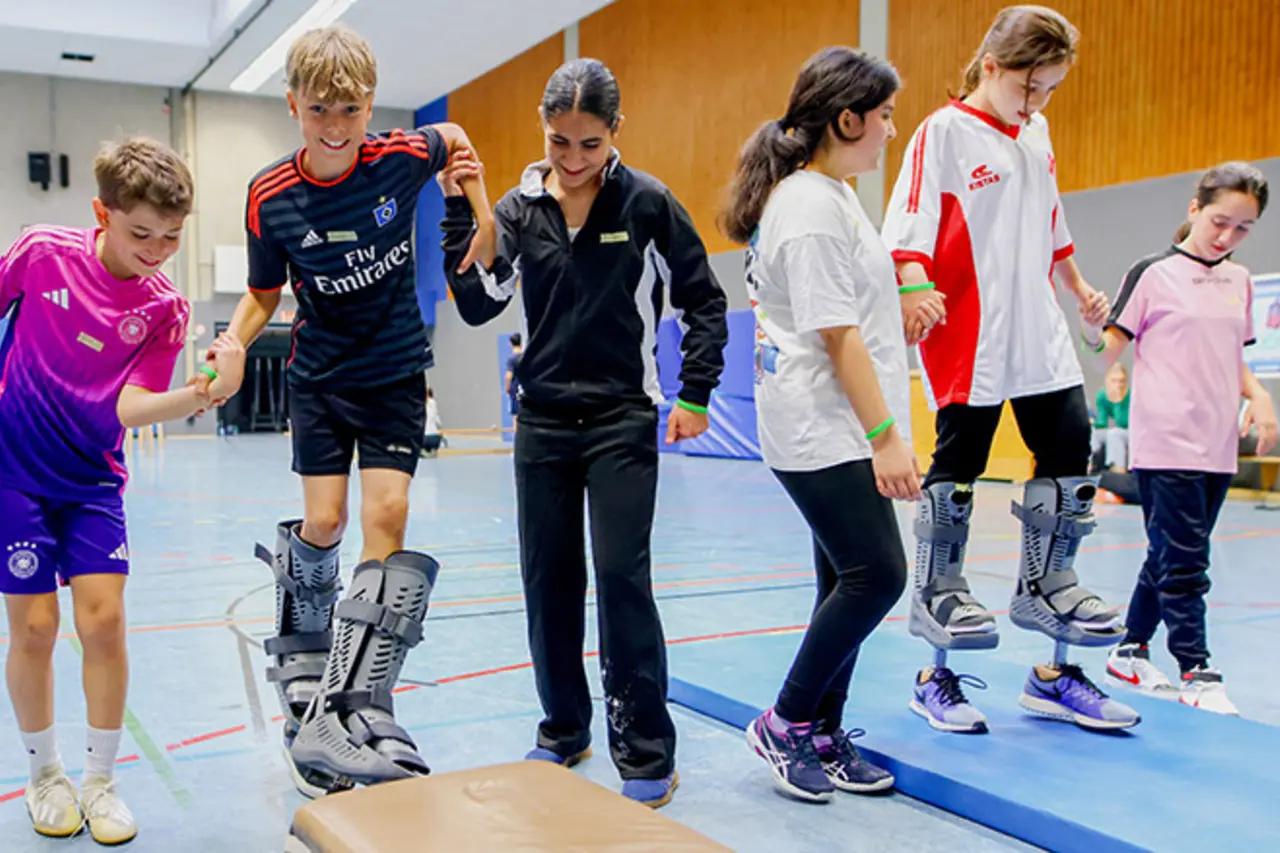 Ein Kind trägt Beinprothesen und balanciert auf einem Kasten. Andere Kinder unterstützen es dabei.