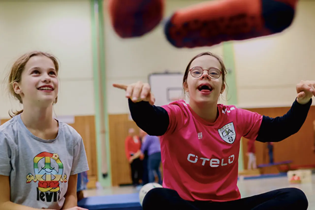 In einer Sporthalle. Zwei lachende Kinder mit und ohne Behinderung machen gemeinsam eine Übung.