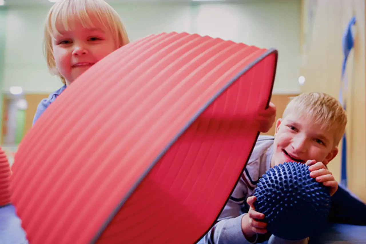 Im Geschicklichkeitsparcours kann man auf bunten Elementen balancieren. Zwei Kinder lachen in die Kamera.