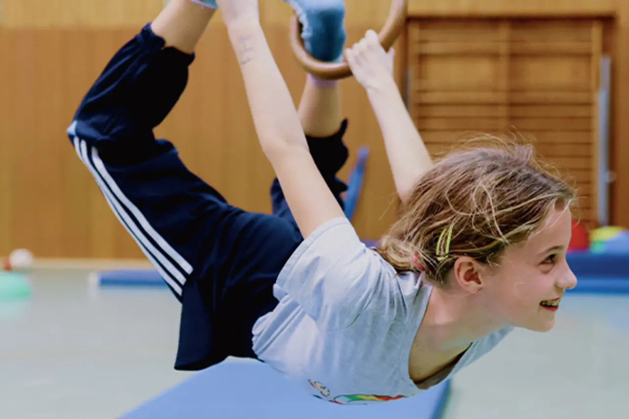 In einer Sporthalle. Ein Kind turnt lachend an den Ringen.