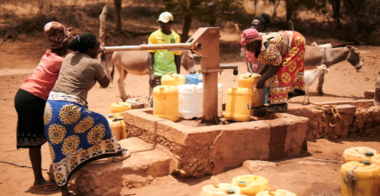 Mehrere Menschen schöpfen Wasser aus einer Handpumpe im Freien und füllen es in gelbe Kanister, während Esel im Hintergrund stehen.