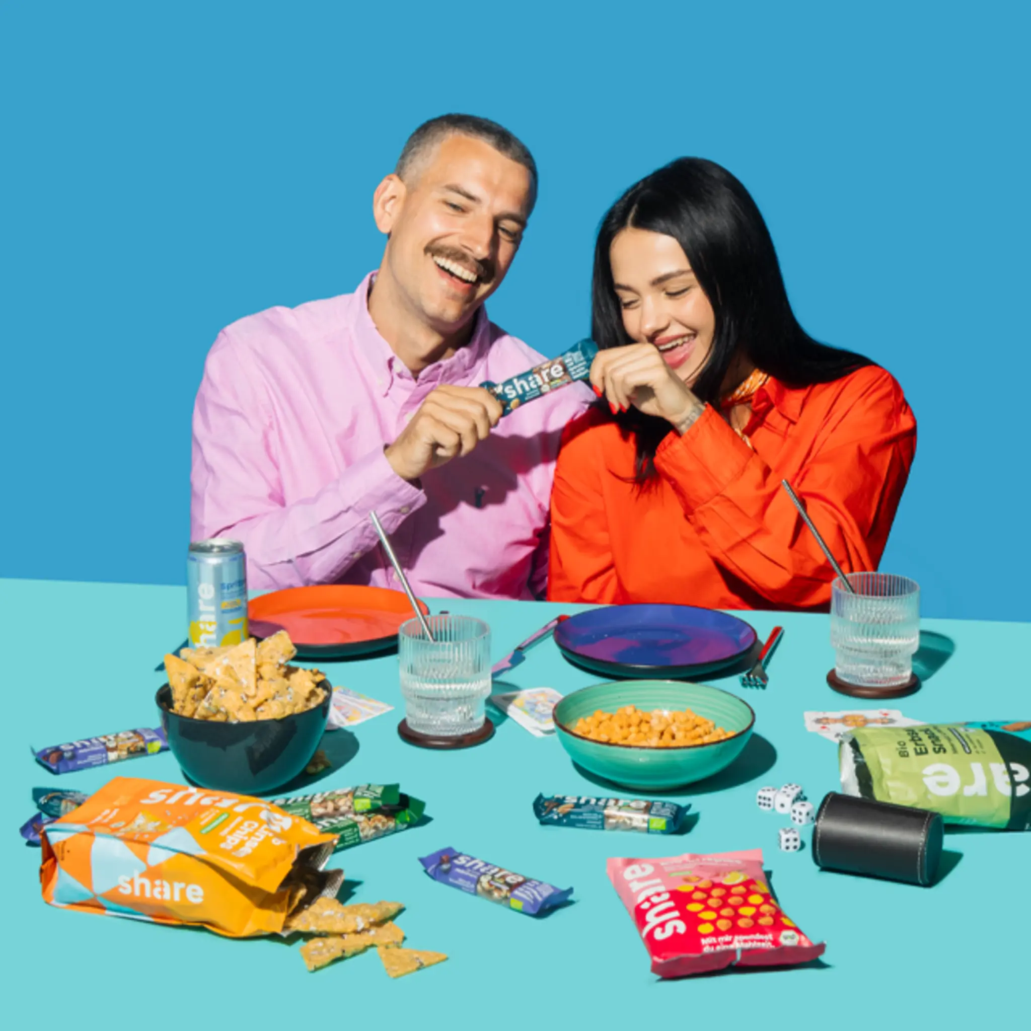 Zwei Personen sitzen an einem Tisch mit Share Snacks und Getränken und haben gemeinsam Spaß beim Essen. 