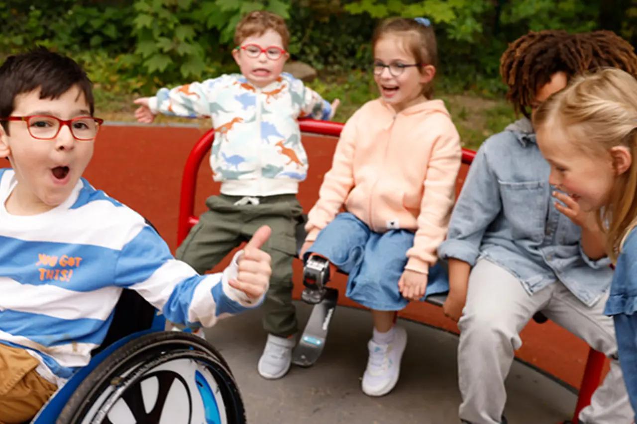 Kinder mit und ohne Behinderung auf einem inklusiven Karussell. In der Mitte sitzt ein Kind im Rollstuhl und zeigt „Daumen hoch“. 
