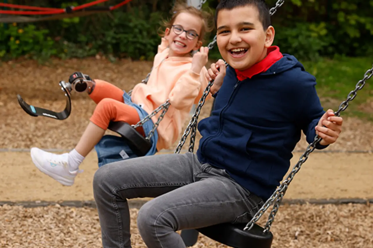 Zwei Kinder sitzen nebeneinander auf zwei Schaukeln. Eines von ihnen trägt eine Beinprothese. 
