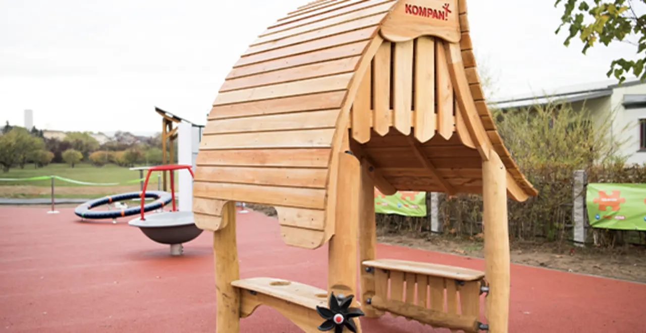 Ein inklusiver Spielplatz mit rotem Gummiboden. Im Vordergrund steht ein inklusives Spielgerät. 