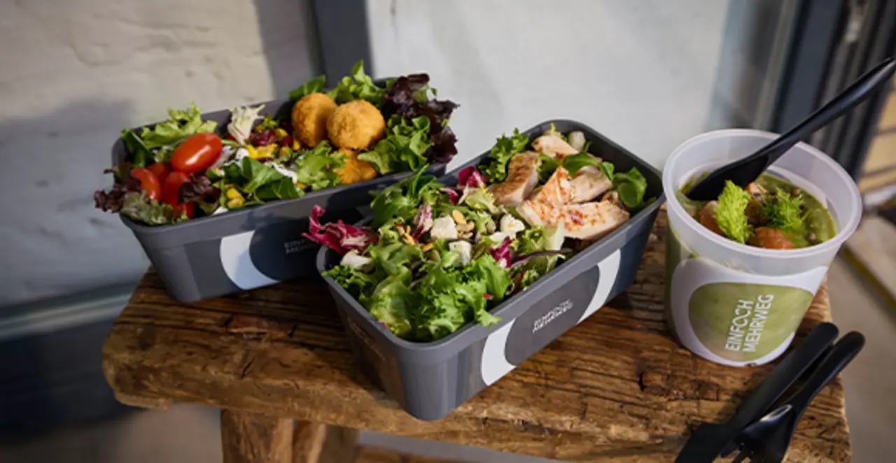 Drei Mehrweg-Pfandbehälter mit frischen Salaten und Suppe stehen auf einem kleinen Holztisch. 