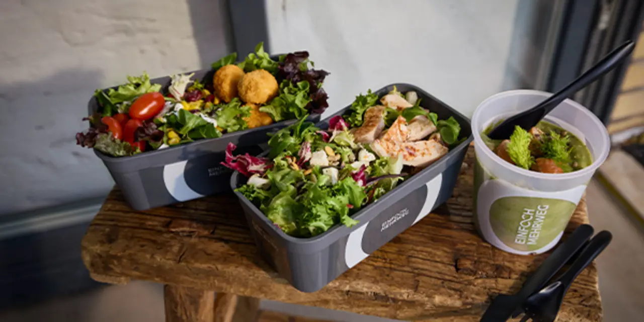 Drei Mehrweg-Pfandbehälter mit frischen Salaten und Suppe stehen auf einem kleinen Holztisch.